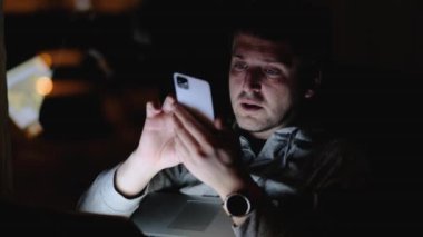 Adam karanlık odada internette cep telefonu ve dizüstü bilgisayarla geziniyor.