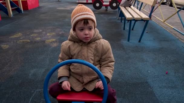 Rapaz bonito em casaco quente senta-se no balanceador no parque infantil — Vídeo de Stock