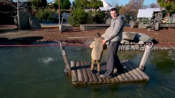 Батько і маленький син перетинають ставок на маленькій дерев'яній платформі — стокове відео