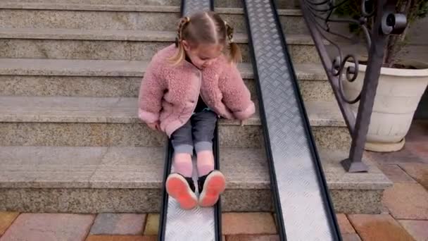 Chica juguetona con trenzas se desliza por la rampa de metal en los escalones — Vídeos de Stock