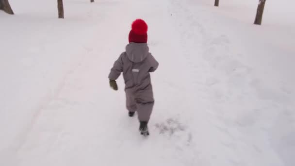 Vidám gyermek szürke kezeslábasban fut végig havas park — Stock videók