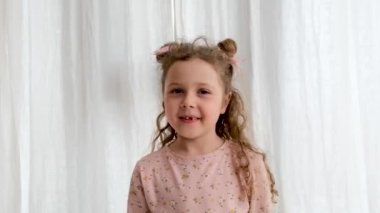 Pembe elbiseli komik küçük kız başını uzay çörekleriyle sallar ve ışık odasındaki beyaz perdelerin yanında duran jesti gösterir.