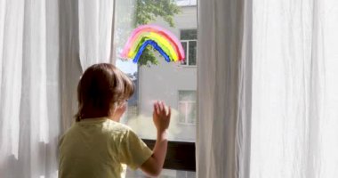 Anaokulu çocuğu renkli gökkuşağıyla pencerenin yanında duruyor