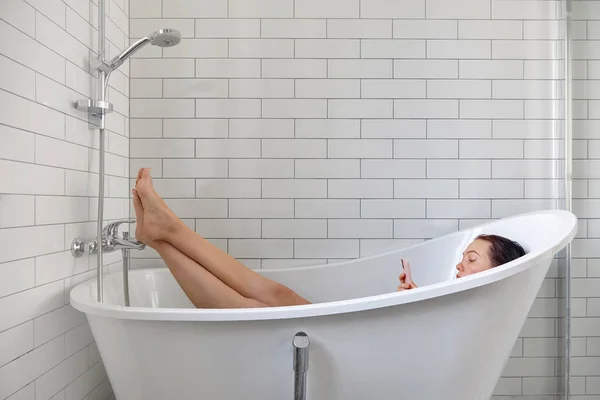 Woman using smartphone in white bathtub side view — 스톡 사진