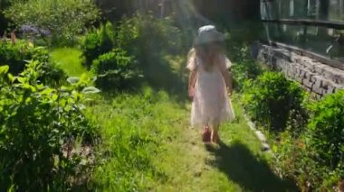 Şapkalı küçük kız yazlık evin yeşil bahçesinde yürüyor.