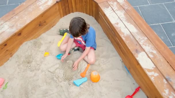 Мальчик в футболке строит башню, сидя в песчаной яме — стоковое видео