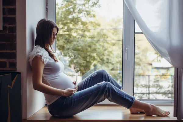 Menina morena grávida sentada no peitoril da janela — Fotografia de Stock
