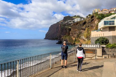 Turistler, Portekiz 'in Madeira kentindeki Camara de Lobos' ta okyanus ve dağlarla manzaranın fotoğrafını çekiyorlar.