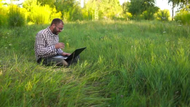 快乐的年轻人在眼镜坐在草地上，在笔记本上打字 — 图库视频影像