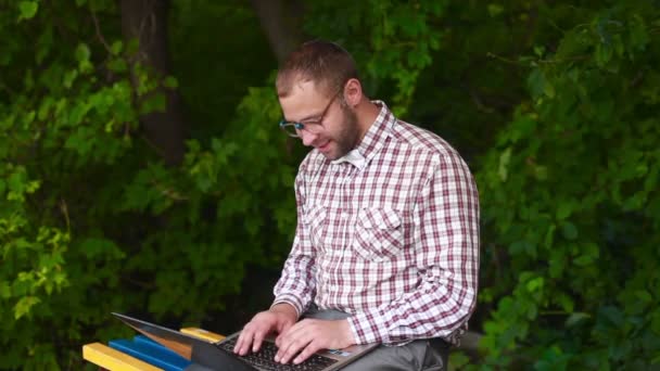 Young man in glasses sitting on a park bench and typing — Stock Video