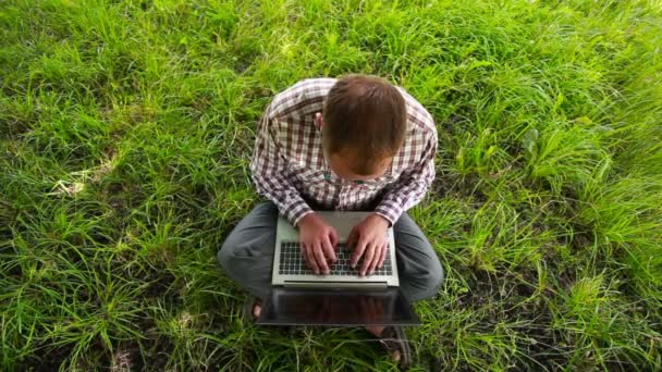 Młody człowiek w okularach, siedząc na trawie i pisania na laptopa — Wideo stockowe