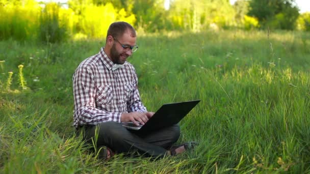 Молодой человек в очках сидит на лужайке и печатает на ноутбуке — стоковое видео