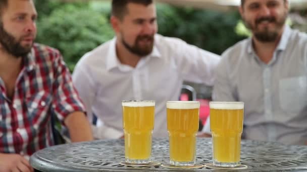 Drie mannen zitten aan de tafel glazen bier en praten — Stockvideo