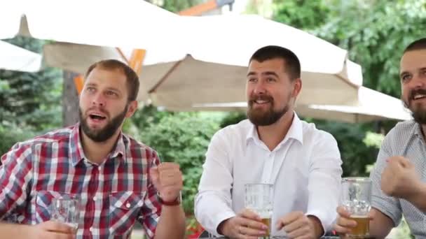 Tres hombres sentados en la cafetería al aire libre viendo el partido y luciendo muy felices — Vídeos de Stock