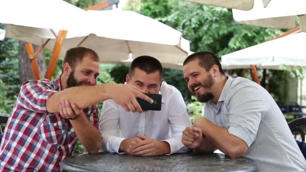 Трое мужчин сидят в кафе, смотрят видео по телефону и смеются — стоковое видео