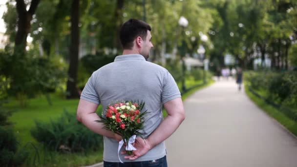 Young man holding lovely bouquet behind his back in park — Stock Video