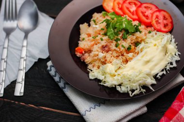 Dilimlenmiş domatesli ve siyah ahşap masada servis edilen sebze salatalı siyah tabakta kızarmış yumurta pilavı..
