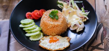 Kızarmış pirinç, sahanda yumurta, salatalık, dilimlenmiş domates, yeşil salata ve garnitür olarak siyah tabağa konmuş kızarmış tavuk.