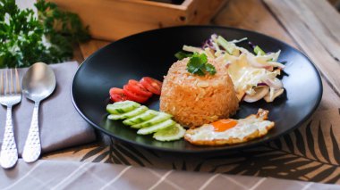 Kızarmış pirinç, sahanda yumurta, salatalık, dilimlenmiş domates, yeşil salata ve garnitür olarak siyah tabağa konmuş kızarmış tavuk.