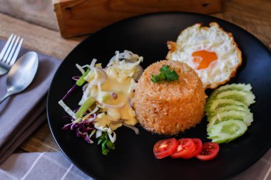 Kızarmış pirinç, sahanda yumurta, salatalık, dilimlenmiş domates, yeşil salata ve garnitür olarak siyah tabağa konmuş kızarmış tavuk.