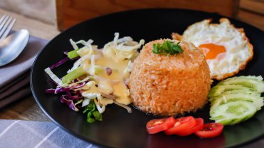 Kızarmış pirinç, sahanda yumurta, salatalık, dilimlenmiş domates, yeşil salata ve garnitür olarak siyah tabağa konmuş kızarmış tavuk.