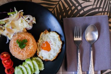 Kızarmış pirinç, sahanda yumurta, salatalık, dilimlenmiş domates, yeşil salata ve garnitür olarak siyah tabağa konmuş kızarmış tavuk.