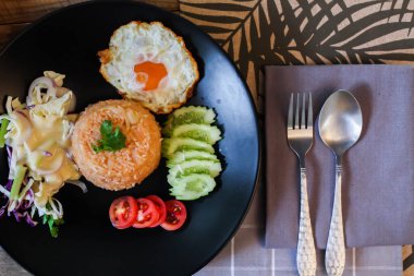 Kızarmış pirinç, sahanda yumurta, salatalık, dilimlenmiş domates, yeşil salata ve garnitür olarak siyah tabağa konmuş kızarmış tavuk.