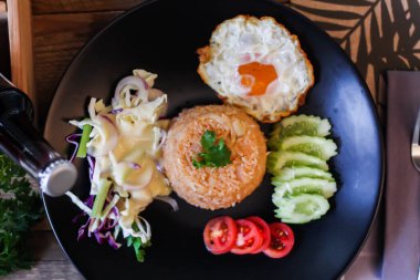 Kızarmış pirinç, sahanda yumurta, salatalık, dilimlenmiş domates, yeşil salata ve garnitür olarak siyah tabağa konmuş kızarmış tavuk.