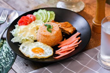 Kızarmış pirinç, sahanda yumurta, salatalık, dilimlenmiş domates, yeşil salata ve garnitür olarak siyah tabağa konmuş kızarmış tavuk..