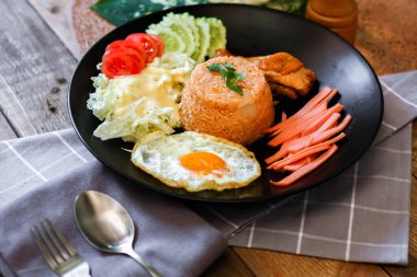 Kızarmış pirinç, sahanda yumurta, salatalık, dilimlenmiş domates, yeşil salata ve garnitür olarak siyah tabağa konmuş kızarmış tavuk..