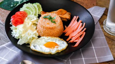 Kızarmış pirinç, sahanda yumurta, salatalık, dilimlenmiş domates, yeşil salata ve garnitür olarak siyah tabağa konmuş kızarmış tavuk..