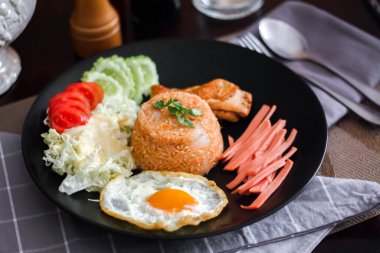 Kızarmış pirinç, sahanda yumurta, salatalık, dilimlenmiş domates, yeşil salata ve garnitür olarak siyah tabağa konmuş kızarmış tavuk..