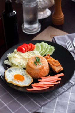 Kızarmış pirinç, sahanda yumurta, salatalık, dilimlenmiş domates, yeşil salata ve garnitür olarak siyah tabağa konmuş kızarmış tavuk..