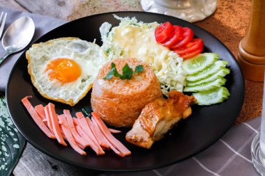Kızarmış pirinç, sahanda yumurta, salatalık, dilimlenmiş domates, yeşil salata ve garnitür olarak siyah tabağa konmuş kızarmış tavuk..