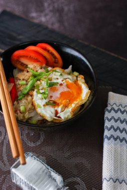 Japon usulü kızarmış pirinç, kızarmış yumurta ve kırmızı domates dilimleriyle birlikte siyah bir kasede servis ediliyor..