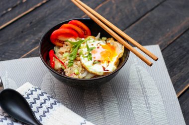 Japon usulü kızarmış pirinç, kızarmış yumurta ve kırmızı domates dilimleriyle birlikte siyah bir kasede servis ediliyor..