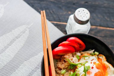 Japon usulü kızarmış pirinç, kızarmış yumurta ve kırmızı domates dilimleriyle birlikte siyah bir kasede servis ediliyor..