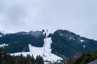 Bir kayak asansörü Megeve 'de yoğun çam ormanlarıyla çevrili kısmen karla kaplı bir yamaçtan yükselir, bulutlu bir kış gökyüzünün altında. Sis ağaçların arasında sürükleniyor, dengesiz bir alp atmosferi yaratıyor..