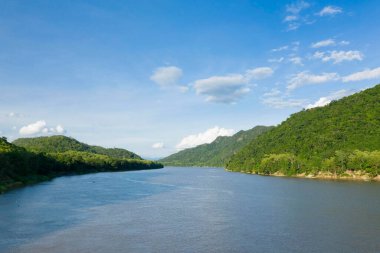 Mekong nehrinin geniş bir manzarası var. Berrak mavi gökyüzünün altında, yemyeşil tepelerle çevrili ve dağınık bulutlar var. Sahne, Güneydoğu Asya 'daki yoğun tropikal bitki örtüsü ve sakin suyu yakalıyor..