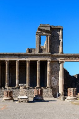 Uzun taş sütunlar ve yıpranmış duvarcılık İtalya 'nın Pompeii kentindeki bazilikanın kalıntılarını oluşturur. Parlak güneş ışığı, canlı mavi gökyüzüne karşı klasik Roma mimarisini ve dokulu yüzeyleri güçlendirir..