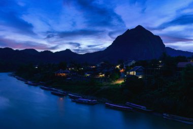 Nong Khiaw, Laos 'un nehir kenarındaki Nong Khiaw köyünü parlak ev ışıkları ve Nam Ou nehri boyunca uzanan ahşap teknelerle kaplar. Çarpıcı kireçtaşı dağları, dönen akşam bulutlarının altında çarpıcı bir siluet oluşturur..