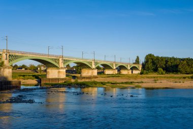 Nevers 'de açık mavi gökyüzünün altında Loire Nehri boyunca uzanan kemerli uzun demir demiryolu köprüsü. Sahnede taş iskeleler, yeşil metal işçiliği, sakin su ve bereketli yaz nehir kıyıları yer alıyor..