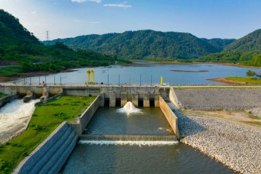 Modern bir hidroelektrik barajı, açık mavi gökyüzünün altındaki gür orman tepeleri ve kayalık setlerle çevrili geniş bir rezervuardan su kanalları oluşturur. Görüntü geometrik yapıları, akan suyu ve mühendislik elementleri arasındaki kontrastı vurgular