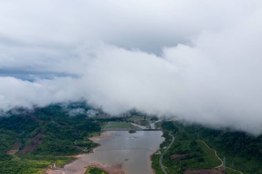 Bereketli yeşil tepelerle çevrili ve yoğun alçak bulutlarla kısmen gizlenmiş kırsal bir rezervuarın havadan bakış açısı. Manzara dolambaçlı yollar, doğal su kanalları ve sisli, sakin bir atmosfere sahiptir..