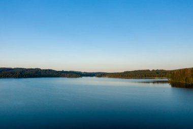 Geniş bir göl, yoğun ormanlarla çevrili sakin mavi su yumuşak gün ışığıyla aydınlatılmış ve engin, bulutsuz bir gökyüzü tarafından çerçevelenmiş..