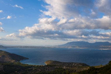 Engebeli tepeler ve uzak dağlarla çevrili derin mavi bir körfez manzarası. Çarpıcı bulutlar dalgalı bir gökyüzünde sürükleniyor. Manzara sükunet ve doğal ihtişam yayar..