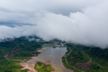 Bereketli yeşil tepelerle çevrili kıvrımlı, çamurlu bir nehrin hava perspektifi ve kısmen alçak bulutlarla kaplanmış. Manzarada doğal su kanalları, yoğun bitki örtüsü ve uzak bir kırsal bölgede sisli bir atmosfer bulunur..