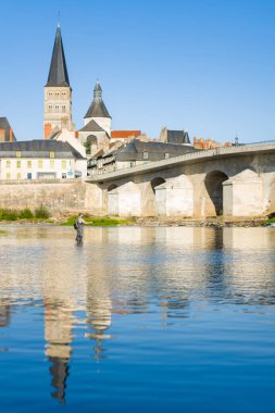 Loire Nehri 'nin sığ sularında, tarihi taş köprüsü ve La Charite sur Loire manastırının nehir kıyısında yükselen kulesiyle yalnız bir balıkçı duruyor. Açık mavi gökyüzü ve sakin yansımalar sakin, resimsel bir sahne oluşturur.