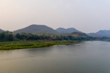Luang Prabang, Laos yakınlarındaki Mekong Nehri 'ni ağaçlık tepeler ve yemyeşil kıyılar üzerinde yumuşak bir sis bulutuyla kaplıyor. Sakin su ve uzak yamaçlar ince doğal tonlarla huzurlu bir nehir manzarası yaratır..