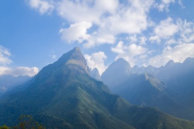 A towering, forested mountain peak rises against a blue sky filled with scattered clouds, with layers of rugged ridges receding into the distance in the Fansipan massif of northern Vietnam.